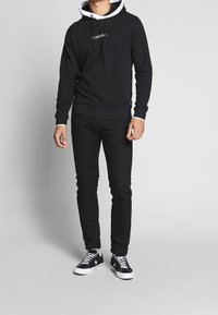 Felpa nera con cappuccio e rivestimento interno bianco, con logo frontale. Indossata con jeans skinny neri e sneakers nere con suole bianche.