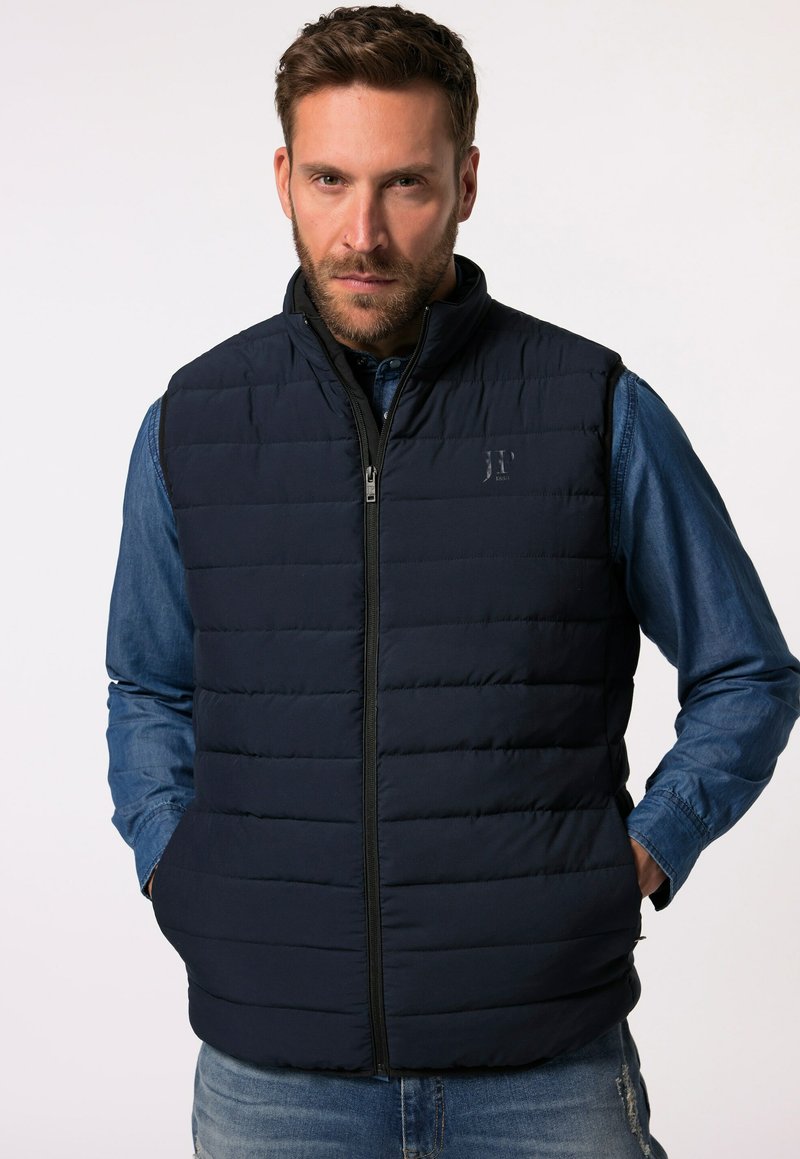 Marineblaue Steppweste mit vertikalem Steppmuster, hochgeschlossenem Kragen, Frontreißverschluss und seitlichen Taschen, getragen über einem Jeanshemd.