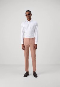 Chemise blanche à boutons, pantalons rose clair et chaussures noires. Les pantalons ont une coupe slim ; des lunettes de soleil ajoutent un look moderne et élégant.