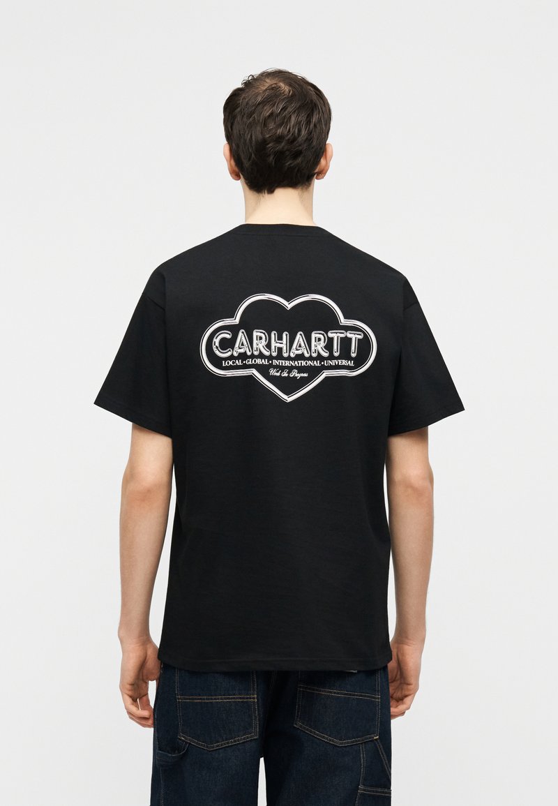 Persona che indossa una maglietta nera Carhartt con logo bianco a forma di nuvola e testo sul retro, abbinata a jeans blu scuro, di spalle su sfondo bianco.