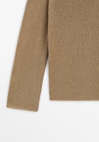 Primo piano della manica e della parte inferiore di un maglione beige a maglia con un motivo a punto spesso e strutturato su sfondo bianco.