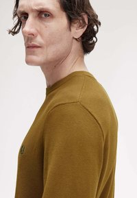 Maglione giallo senape con texture, colletto rotondo e logo ricamato verde tenue al centro, che evidenzia uno stile aderente.