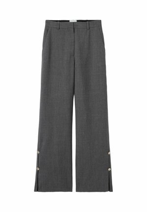 Mujer de pie con una chaqueta gris corta, pantalones anchos a juego y zapatos marrones sobre un fondo liso y claro.