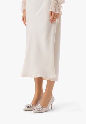 Kobieta w kremowej spódnicy midi i jasnoszarych szpilkach zdobionych kwiatami, stojąca z rękami spoczywającymi obok spódnicy.