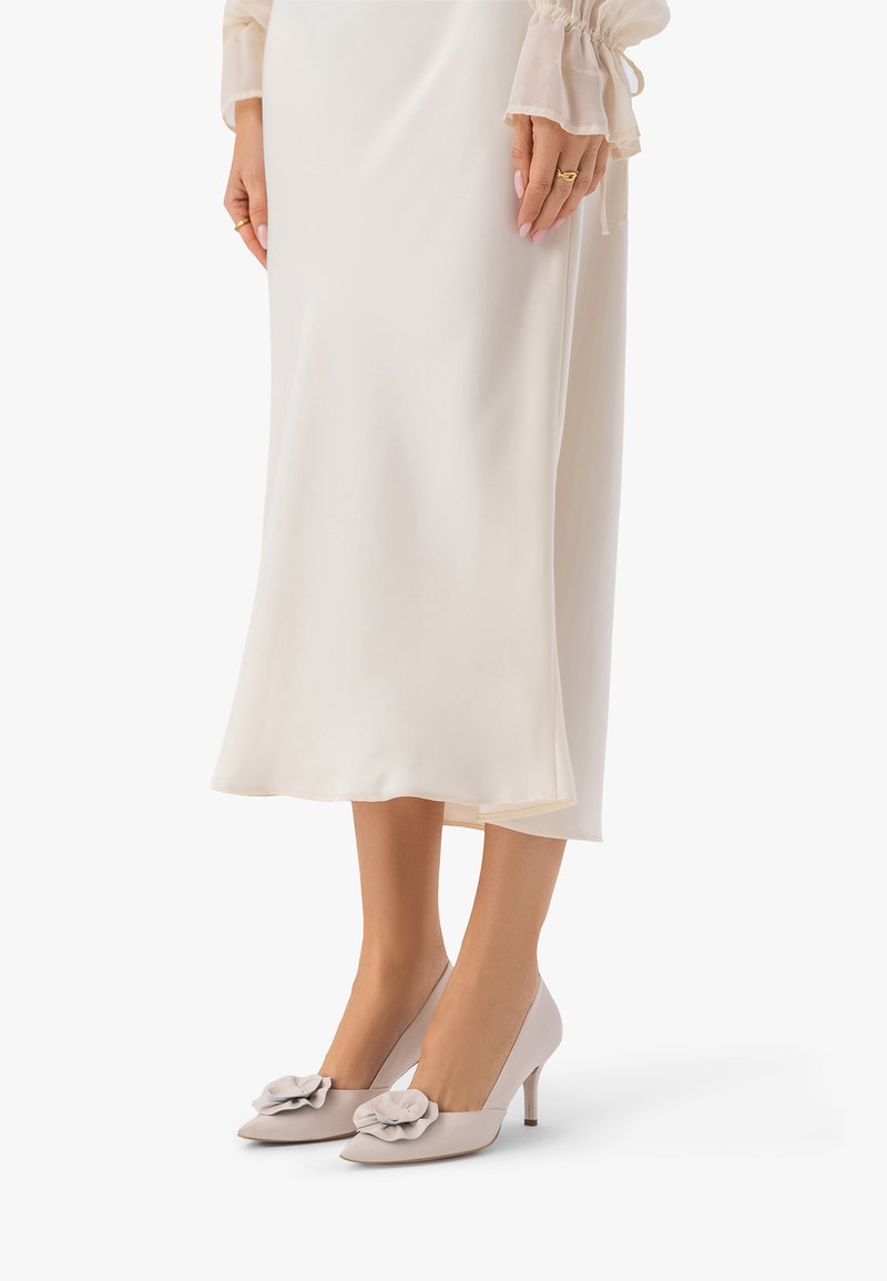 Femeie purtând o fustă midi crem și pantofi cu toc înalt gri deschis cu decor floral, stând cu mâinile pe lângă fustă.