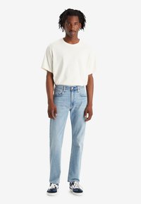 Uomo con capelli intrecciati che indossa una maglietta bianca semplice, jeans azzurri chiari e sneakers blu e bianche, in piedi davanti a uno sfondo neutro.