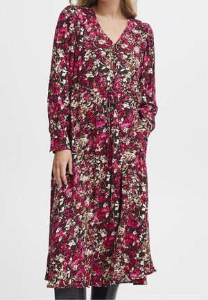 Frau trägt ein langärmeliges, geblümtes Kleid mit pinken, violetten, weißen und braunen Blumen und einem Bindegürtel in der Taille.