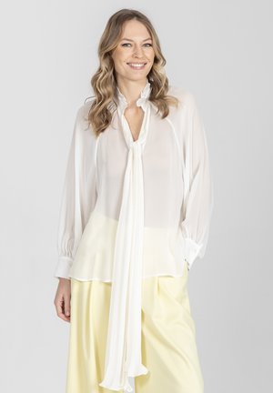 Uśmiechnięta kobieta o falowanych blond włosach, ubrana w białą bluzkę z długim rękawem i wiązaniem przy szyi oraz jasnżółte szerokie spodnie, na szarym tle.