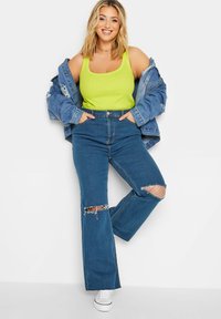 Top verde chiaro, jeans a vita alta in denim blu sfilacciati con orli strappati e una giacca in denim blu. Sneakers bianche completano l'outfit.