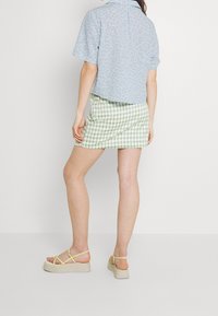 Camicia blu a maniche corte con motivo floreale, mini gonna verde e bianca a scacchi, sandali gialli con cinturini e suola a piattaforma. Vista da dietro.