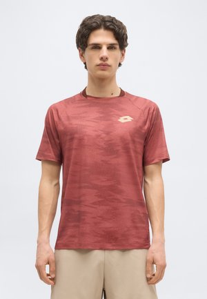 Fiatal férfi rozsdavörös mintás, rövid ujjú sportpólóban és bézs rövidnadrágban, egy egyszerű fehér háttér előtt áll.