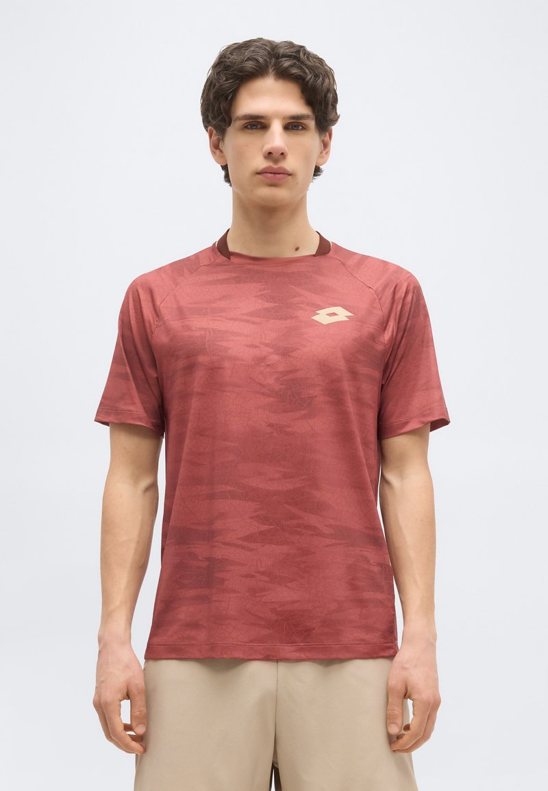 Νεαρός άνδρας που φοράει αθλητική μπλούζα κοντομάνικη με σχέδια σε σκουρόχρωμο κόκκινο και μπεζ σορτς, στέκεται μπροστά σε απλό λευκό φόντο.