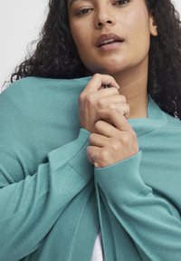 Turkossfärgad stickad cardigan med en mjuk textur, med en bred krage och ribbade ärmslut, omsvept runt axlarna.