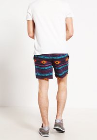 Bañador de hombre con un patrón multicolor que presenta diseños geométricos en rojo, amarillo y azul sobre un fondo oscuro, con un ajuste relajado.