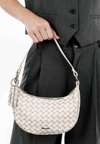 Beige kuukkarin muotoinen laukku, jossa on geometrinen kuvio, hopeiset yksityiskohdat ja irrotettava kortti, pidettynä kädessä vaaleanharmaan asun kanssa.