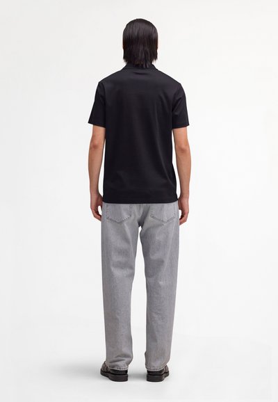 Person facing away wearing a black short-sleeve shirt, light gray jeans, and black shoes on a plain white background.