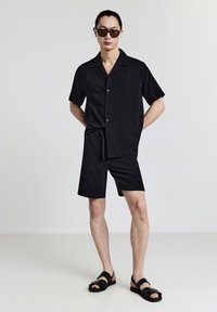 Maglietta nera a maniche corte e pantaloni corti coordinati con un motivo a diamante testurizzato. Indossati con sandali neri e occhiali da sole oversize.