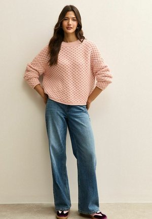 Hellrosa strukturierter Pullover mit weitem Rundhalsausschnitt und hochgekrempelten Bündchen, kombiniert mit locker sitzenden blauen Jeans und dunkelvioletten Schuhen.