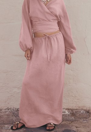 Donna che indossa un lungo top rosa avvolgente e una gonna coordinata con coulisse, con sandali neri, in piedi su un pavimento in pietra contro un muro bianco.