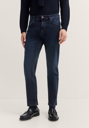 Man wearing dark blue slim-fit jeans, black loafers, and a black sweater with a scarf against a plain background.