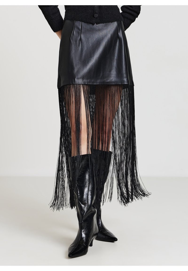 Mini-jupe en cuir noir avec un overlay à longues franges, associée à des bottes noires brillantes montantes jusqu'au genou avec un bout pointu, sur un fond clair.