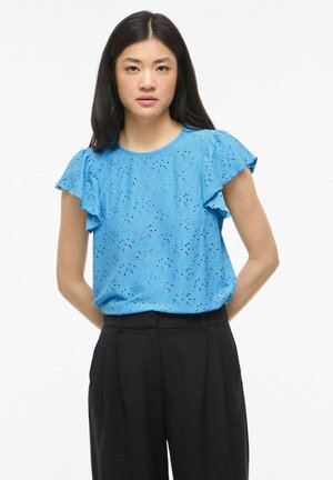 Femme aux longs cheveux foncés portant un chemisier bleu à manches courtes avec œillets et un pantalon noir taille haute, debout les mains derrière le dos.