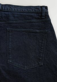 Poche arrière d'un jean en denim bleu foncé avec couture visible et passants de ceinture sur un fond clair.