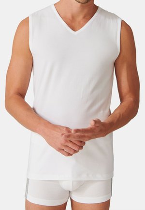 Mann, der ein schlichtes weißes ärmelloses V-Ausschnitt-Shirt und weiße Boxershorts trägt, steht mit gefalteten Händen vor einem neutralen Hintergrund.