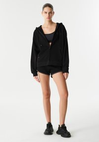 Czarny zapinany na zamek hoodie z troczkami, w połączeniu z czarnym stanikiem sportowym i czarnymi szortami. Model nosi czarne sneakersy, prezentując sportowy strój.