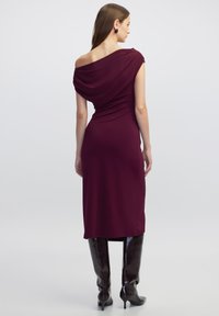 Burgunderfarbenes Midikleid mit schulterfreiem Drapé-Design, figurbetonender Silhouette und weicher Textur. Kombiniert mit hohen, glänzenden schwarzen Stiefeln.