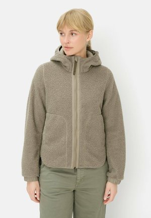 Kvinde iført en lysebrun fleecejakke med lynlås, hætte og elastiske manchetter, kombineret med olivengrønne bukser, som kigger let mod venstre.