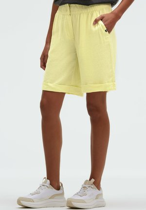 Person trägt hellgelbe knielange Shorts mit aufgerollten Säumen und weiße Turnschuhe, steht mit einer Hand in der Tasche.