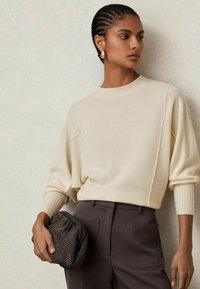 Žena s vrkočmi, oblečená v krémovom svetri a tmavých nohaviciach, drží štruktúrovaný čierny clutch a pozerá sa do strany na neutrálnom pozadí.