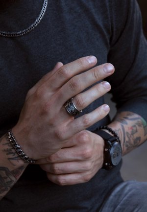Silver chain necklace and a dark-colored, textured shirt. The hand wears a detailed black ring and a beaded bracelet, with visible tattoos.
