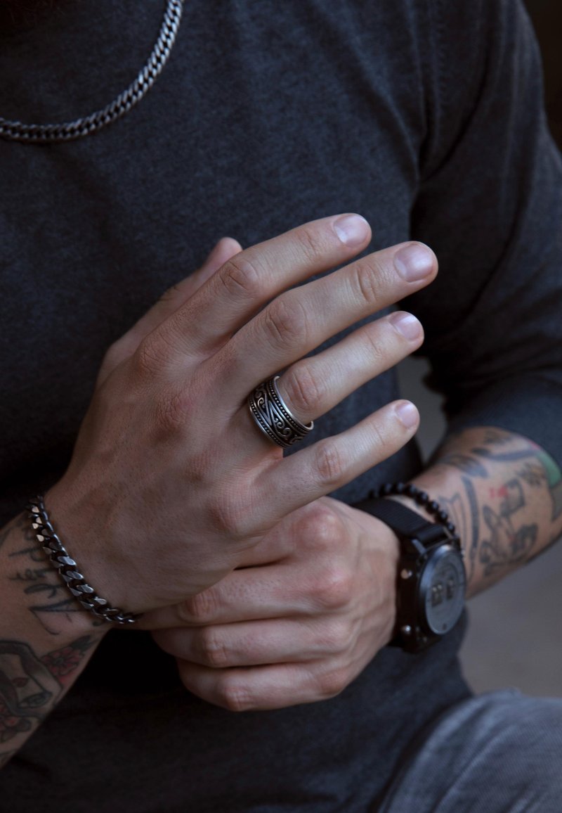 Zilveren ketting en een donkergekleurde, gestructureerde shirt. De hand draagt een gedetailleerde zwarte ring en een kralenarmband, met zichtbare tatoeages.