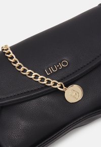 Borsa a mano in pelle nera con tracolla in catena dorata e charm. Presenta il nome del marchio in rilievo e una superficie texturizzata. Design compatto e rettangolare.