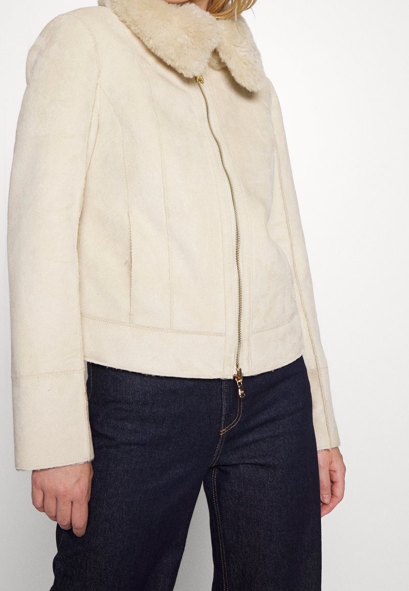 Giacca in finta shearling beige con colletto morbido, chiusura con zip e tasche laterali. Presenta un design cropped e una texture liscia.