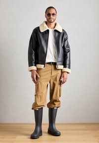 Man wearing black leather jacket with beige shearling collar and cuffs, white shirt, tan cargo pants, black rain boots, and sunglasses standing indoors.