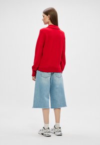 Červený pletený sveter s širokým golierom, kombinovaný s svetlomodrými denimovými culotami, so surovým lemom a uvoľneným strihom. Biele tenisky s čiernymi akcentmi.