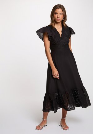 Femme portant une robe midi noire avec des manches volantes, des détails en dentelle sur le corsage et l'ourlet, et des sandales plates argentées à brides, debout devant un fond blanc.