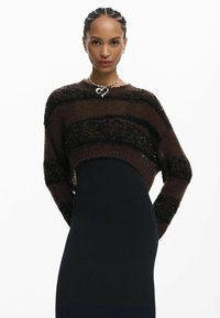 Fonott hajú nő barna és fekete csíkos kivágott pulóvert visel fekete ruha fölött, ezüst szív alakú nyaklánccal kiegészítve.