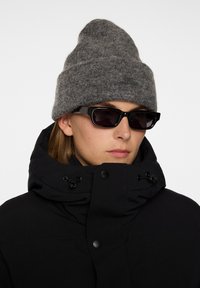 Musta puffatakki korkean kauluksen kanssa, harmaa villapipo ja kulmikkaat mustat aurinkolasit, esittelee sileitä pintoja ja kontrastisia värejä.