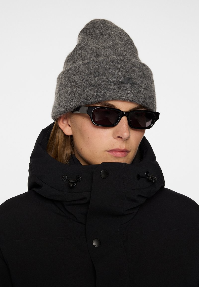 Musta puffatakki korkean kauluksen kanssa, harmaa villapipo ja kulmikkaat mustat aurinkolasit, esittelee sileitä pintoja ja kontrastisia värejä.