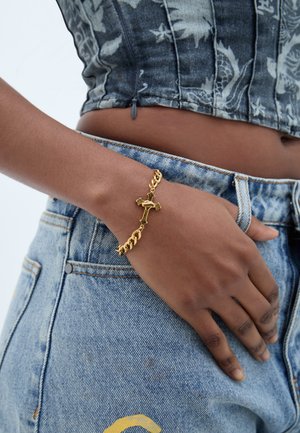 Κοντινό πλάνο ενός ατόμου που φοράει βραχιόλι με χρυσό σταυρό στον καρπό, με τζιν παντελόνι και μοτίβο κορυφαίου τοπ να φαίνονται.