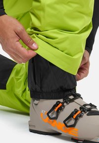 Helles grünes wasserdichtes Hosen mit schwarzen Bündchen, elastischen Öffnungen und einer Hand, die den Saum nahe einem grauen Skischuh mit orangefarbenen Akzenten anpasst.