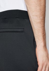 Černé sportovní kalhoty s texturovaným povrchem, s žebrovaným pasem, zadní kapsou a slim fit střihem. Materiál vypadá pružně.