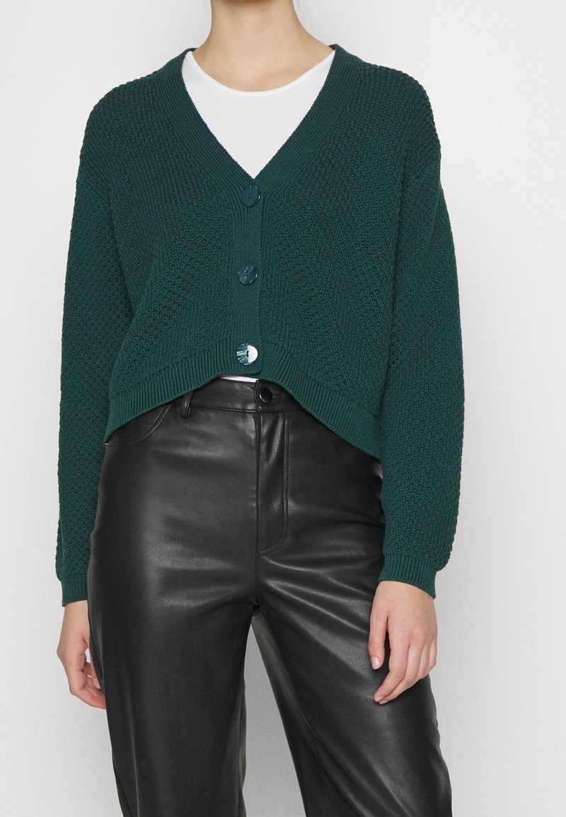 Personne portant un cardigan boutonné texturé vert foncé par-dessus un haut blanc, associé à un pantalon en cuir noir taille haute.