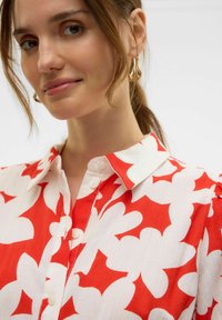 Camicia a fiori rossi e bianchi con colletto, bottoni e maniche corte. Realizzata in tessuto leggero con una superficie testurizzata.