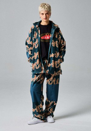 Persona con pelo rubio corto que lleva una chaqueta y pantalones azules a juego con un patrón de llamas naranjas y zapatillas blancas.