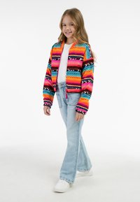 Bunte, gestrickte Jacke mit horizontalen Streifen in Pink, Orange und Blau, kombiniert mit hellen, ausgestellten Jeans und weißen Sneakers.